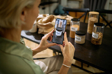 Caucasian middle aged woman using smartphone to photograph labeled glass jars filled with grains and seeds on wooden table, hands and partial profile visible, kitchen setting
