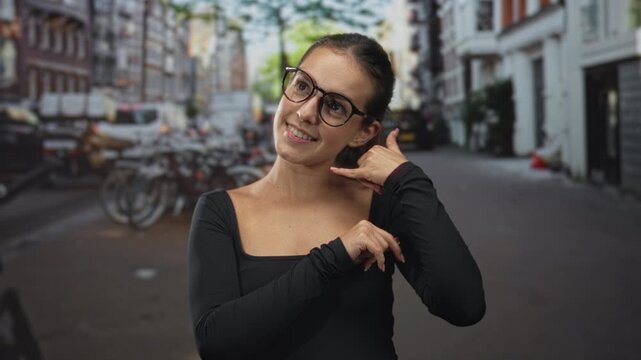 Woman with glasses making call me hand gesture on a city street lined with bicycles and brick buildings wearing a black long sleeve shirt; playful flirtation.