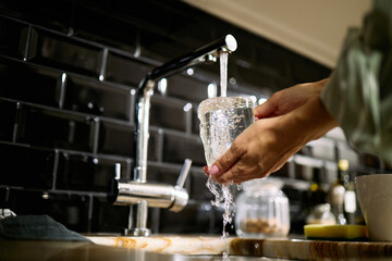 Caucasian adult woman washing glass under running water at kitchen sink, hands holding transparent cup with water splashing, black tiled backsplash visible in background