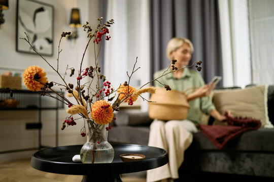 Middle aged Caucasian woman sitting on sofa using smartphone with blurred focus while foreground showing vase with autumn flowers and berries on round table - Powered by Adobe