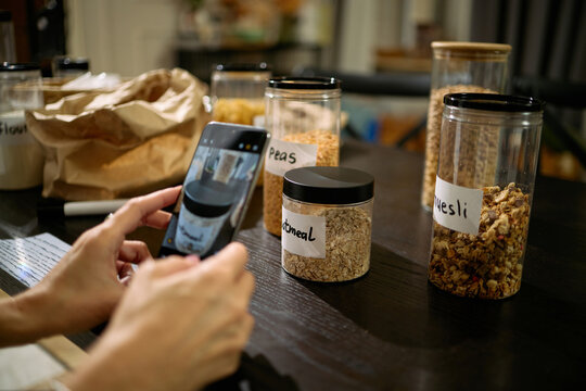 Caucasian young adult woman photographing labeled food storage jars with smartphone on kitchen table, focusing on containers filled with oatmeal, peas, and muesli for organization