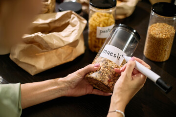 Caucasian middle aged woman labeling transparent food container with marker, organizing dry breakfast cereal on dark table with other jars and paper bag visible in background