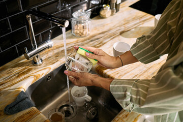 Caucasian woman washing glass under running water in kitchen sink, holding sponge and cleaning dishware, visible hands and arms showing adult female engaged in household chore