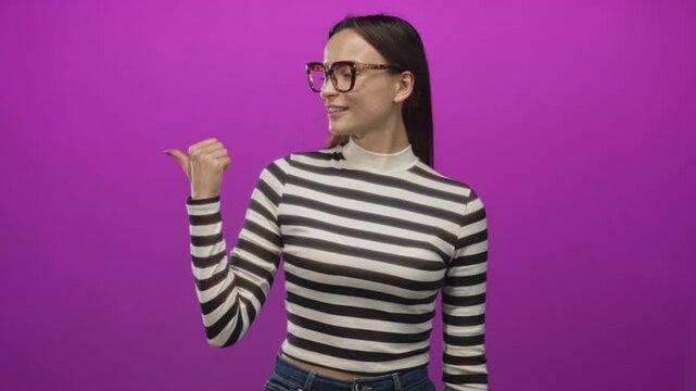 Woman wearing tortoiseshell glasses points her thumb to the left in a purple studio; confidence optimism.