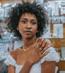 Chest-high portrait of a young woman with curly hair, a diamond necklace and a green gem ring, staring at the camera in a jewelry store.