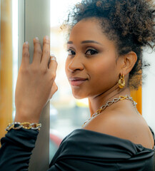 Portrait of an elegant young woman with curly hair, showing a set of thick chain jewelry, while leaning against a window.