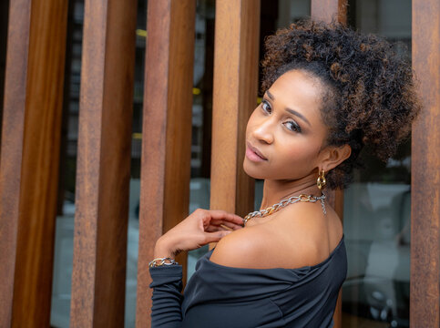 Young African-American woman, elegant with curly hair, dressed in black, posing and showing a necklace and thick chain earrings of modern and striking style.