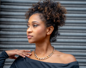 Profile portrait of an elegant young woman with curly hair and a bimetallic chain necklace, touching her shoulder and looking thoughtfully to the side.