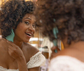 Elegant young woman with curly hair, looking in the mirror and smiling. She is trying on a gold necklace and a light blouse, evaluating her appearance.