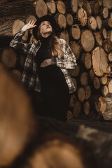 Pregnancy and Nature: An elegant pregnant woman, in a stylish hat and a long black dress, posing against a pile of wooden logs in the warm light of the autumn sun