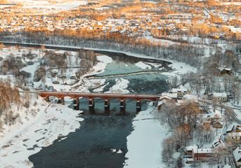 Aerial winter view of historic red brick bridge over partly frozen river, snowy trees glowing in golden light and small town houses in background, scenic European landscape