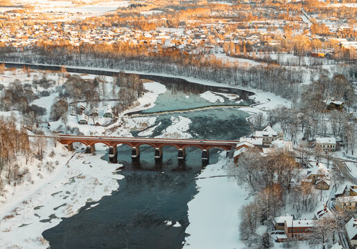 Aerial winter view of historic red brick bridge over partly frozen river, snowy trees glowing in golden light and small town houses in background, scenic European landscape