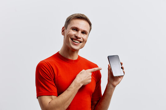 Smiling man in orange t-shirt hold smartphone point finger show screen isolated white background digital communication technology concept