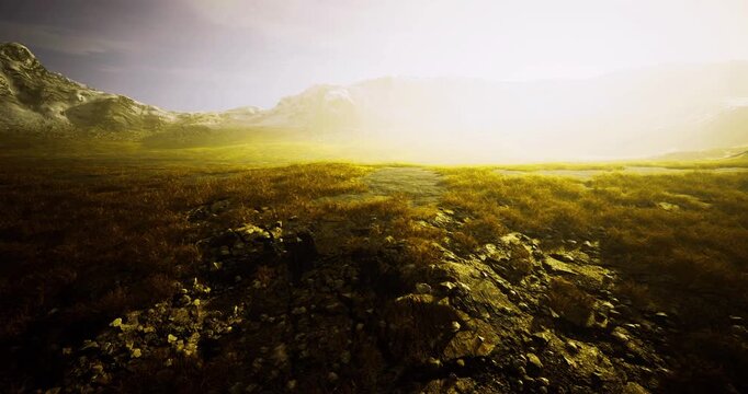 Golden light illuminates a rugged landscape featuring rocky terrain and patches of green grass. Mist settles over distant mountains, creating an ethereal atmosphere at dusk.