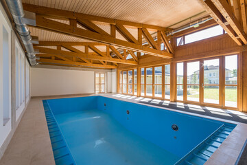 Large indoor swimming pool features a bright atmosphere with wooden beams and ample natural light filtering through expansive windows. Ideal for relaxation and leisure activities at home