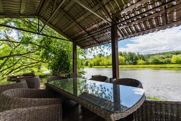 A riverside dining area features a glass table surrounded by wicker chairs. Lush greenery reflects off the calm water while fluffy clouds drift in the blue sky on a sunny day