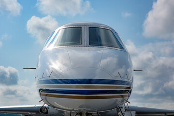 Tight front view of a private jet cockpit beneath a partly cloudy sky. The aircraft's polished nose and dual windshields reflect the environment with clarity.