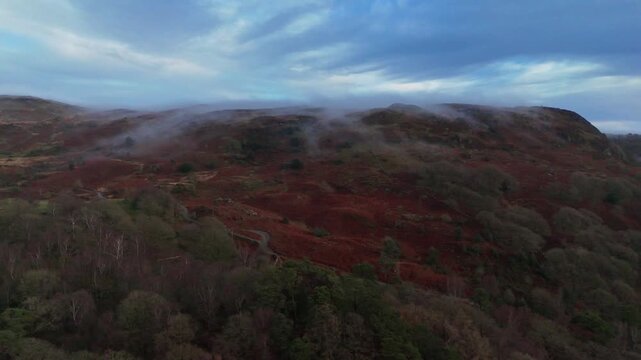 Drone view of mist moving across an autumn hillside with red bracken and woodland beneath a cool, atmospheric sky.
