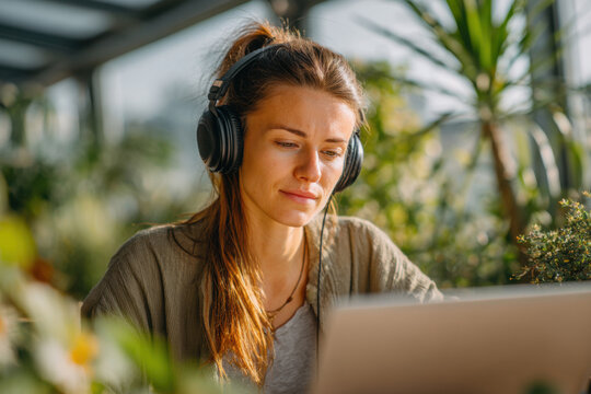 A young businesswoman with a ponytail and headphones on her head sits at her desk in front of a laptop. Blur background with plants. Focused flow