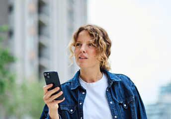 Portrait of a happy young woman tourist travelling or student or a businesswoman entrepreneur, using a smartphone phone or texting outdoors sitting on a bench in park in city