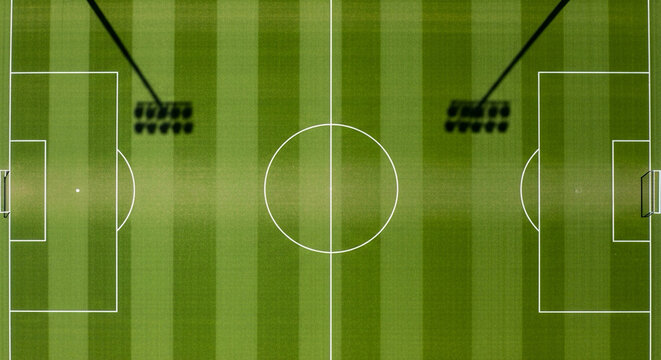 Overhead drone view of a perfectly marked football pitch, empty, sunlight casting shadows of goalposts, symmetrical composition.