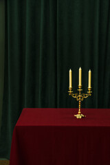 A bronze candlestick with candles stands on a red tablecloth against a background of green curtains.
