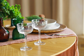 Close up of elegant crystal glasses, plates and flowers on wooden table with green decorations and warm holiday ambiance. Perfect for celebrations, cozy and romantic gatherings, seasonal home decor.
