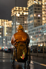 Unrecognizable disabled man in wheelchair with hood on and backpack walking on pathway late at night in rgb color light. Male with disability spending time alone on fresh air to keep activity.