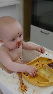 Baby with a messy face sits in a high chair and happily with appetite drink water from a sippy cup. Concept of early childhood, feeding time, and introducing solids to infants. parental care