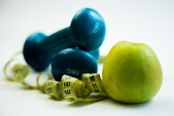 Blue dumbbells and a green apple are positioned alongside a measuring tape, representing fitness and health, encouraging a balanced lifestyle and personal wellness