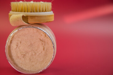 Exfoliating scrub jar with wooden brush on a bright red background, showcasing natural skincare products for beauty routines and self-care practices for wellness lovers