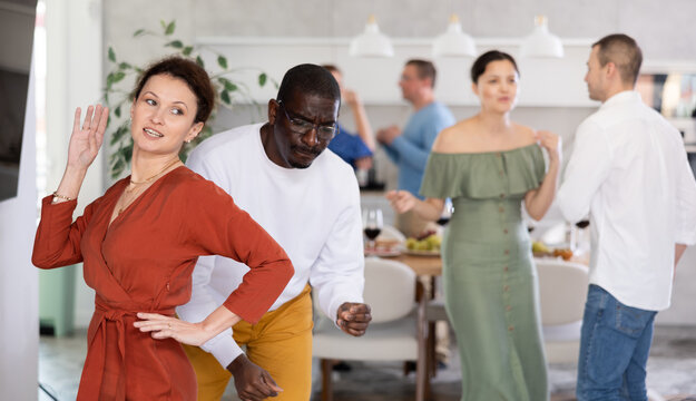 Emotional lively woman in terracotta dress with joyous expression dancing at friendly multicultural house party..