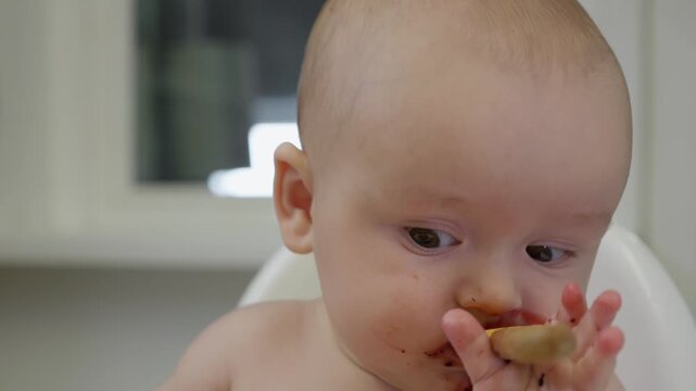 Baby sits in a high chair and happily eating pureed food with a spoon. Emotional messy baby face covered in food. Concept of early childhood, feeding time, and introducing solids to infants