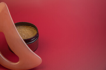 Round jar containing brown sugar scrub is positioned next to a pink applicator on a bright red background, emphasizing beauty and skincare themes with copy space