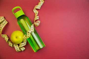 Bright green water bottle alongside a yellow measuring tape and a fresh apple on a vivid red background, representing health and fitness themes for wellness motivation