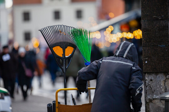 Rake and broom on worker’s cart in busy street scene with hooded figure in foreground and soft festive lights illuminating crowded urban background - Powered by Adobe