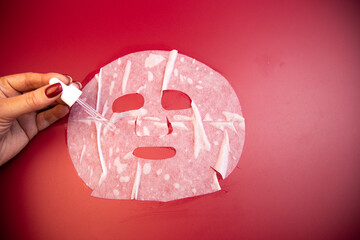 Hand of a woman applying serum to a facial mask against a bold red backdrop, highlighting skincare rituals and the importance of self-care in beauty routines