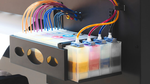 Colored ink cartridges on a large industrial printer, with transparent tubes containing ink connected to the cartridges.