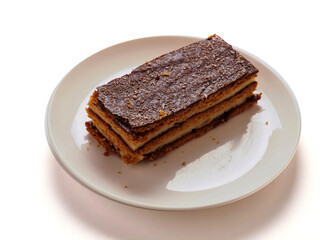 A dessert layered cake with chocolate is on a white plate on white background. The dessert is a cake with a brown crust. The plate is white and the dessert is the main focus of the image