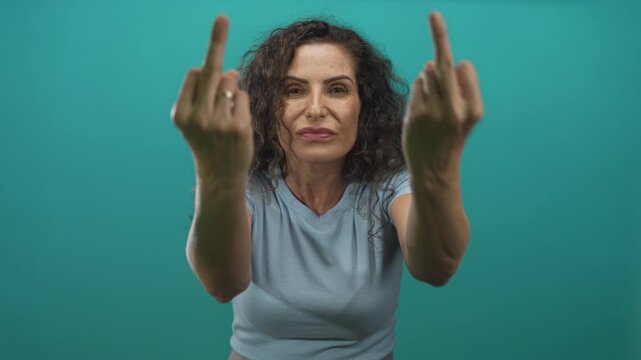 Hispanic woman with curly hair and blue t shirt extends middle fingers in teal studio environment; defiance.