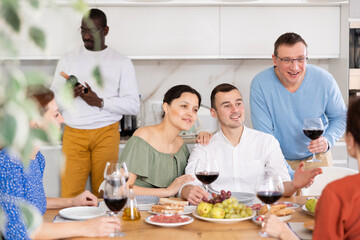 Happy middle-aged men and women sitting at table with a glass of wine while having friendly conversation at home