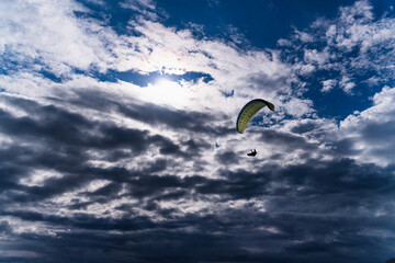 Paraglider soars through dramatic cloudscape as sun breaks through textured sky, capturing freedom, adventure and the thrill of high altitude flight over open landscape