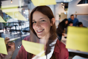 Smiling young woman writing notes on glass board during creative meeting. Focused professional contributing ideas in a modern collaborative workspace. Creativity and teamwork concept.