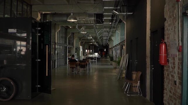 open space modern office old abandoned factory, pipes run under ceiling, large lamps hang, center corridor there are tables, assistant works laptop, partitions offices are made of glass