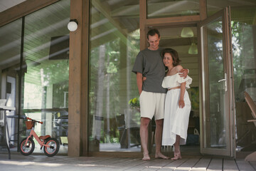 A couple stands together on a wooden porch of a cabin, smiling and embracing each other. The bright...
