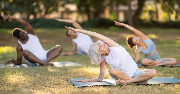 Multinational people of different ages in park practicing yoga. Amateurs performing gymnastic. Senior woman yoga master and followers do padmasana, svastikasana. People practice yoga during in nature