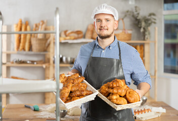 Bakery employee holds basket of ready-made croissant. Male baker holds basket with finished products, shows many different croissants.