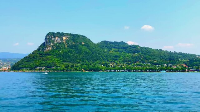 Impressive view from Lago di Garda at Rocca di Garda near Bardolino, the vegetation-topped promontory standing tall, surrounded by smoothly rippling mirroring luminescent lake waters, hills, clear sky