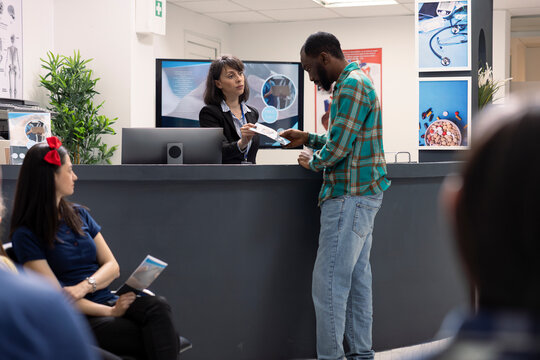 White woman at front desk hands promotional flyer to male outpatient, explaining after treatment options in hospital waiting area. Female receptionist provides health insurance advice to black man.