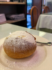 Maritozzo dusted with powdered sugar sits elegantly on a white plate, accompanied by a silver spoon, showcasing a delightful dessert experience in a cozy cafe setting